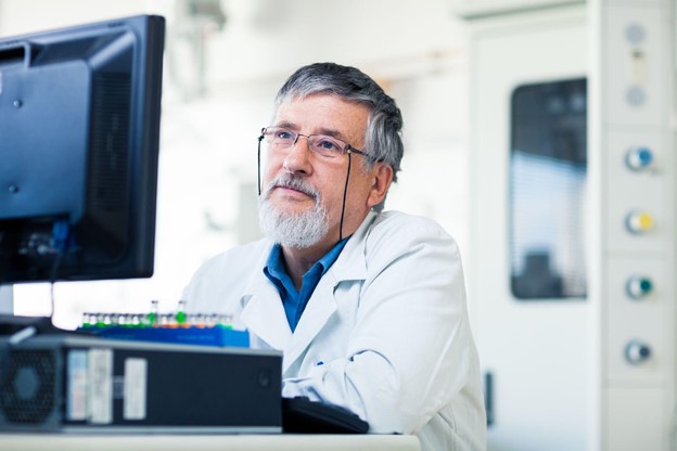 Male researcher in a white labcoat  observing information on a computer screen.